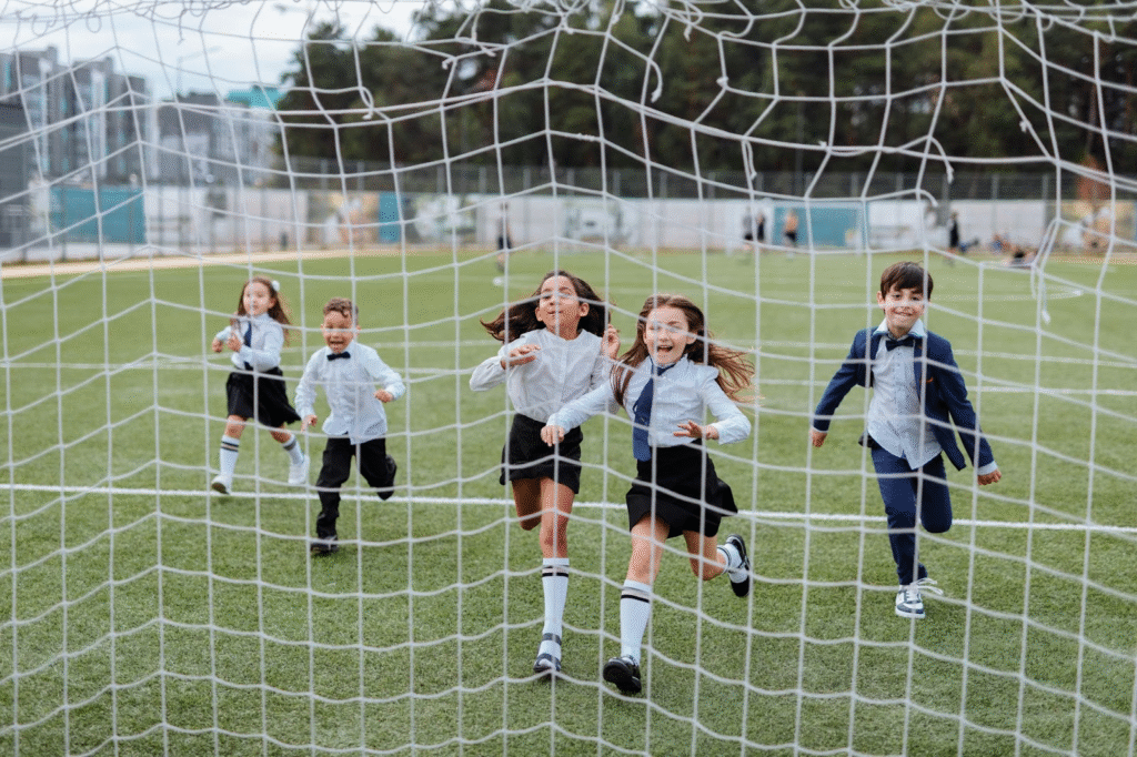 Children in school uniforms joyfully running through a soccer goal net on a field trip.