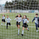 Children in school uniforms joyfully running through a soccer goal net on a field trip.
